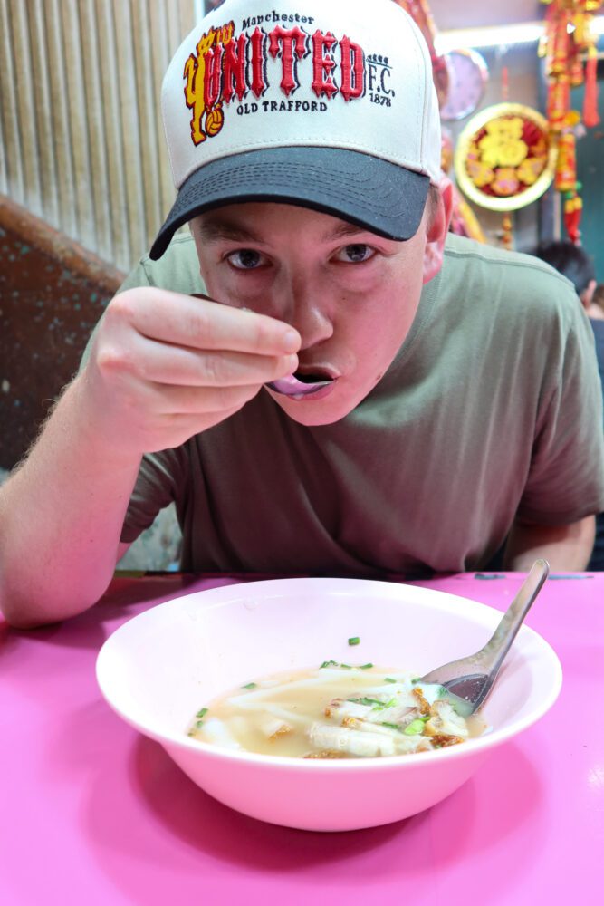 Man enjoying a bowl of rolled noodle soup at a vibrant street food stall in Bangkok’s Chinatown, under colorful Chinese New Year decorations.