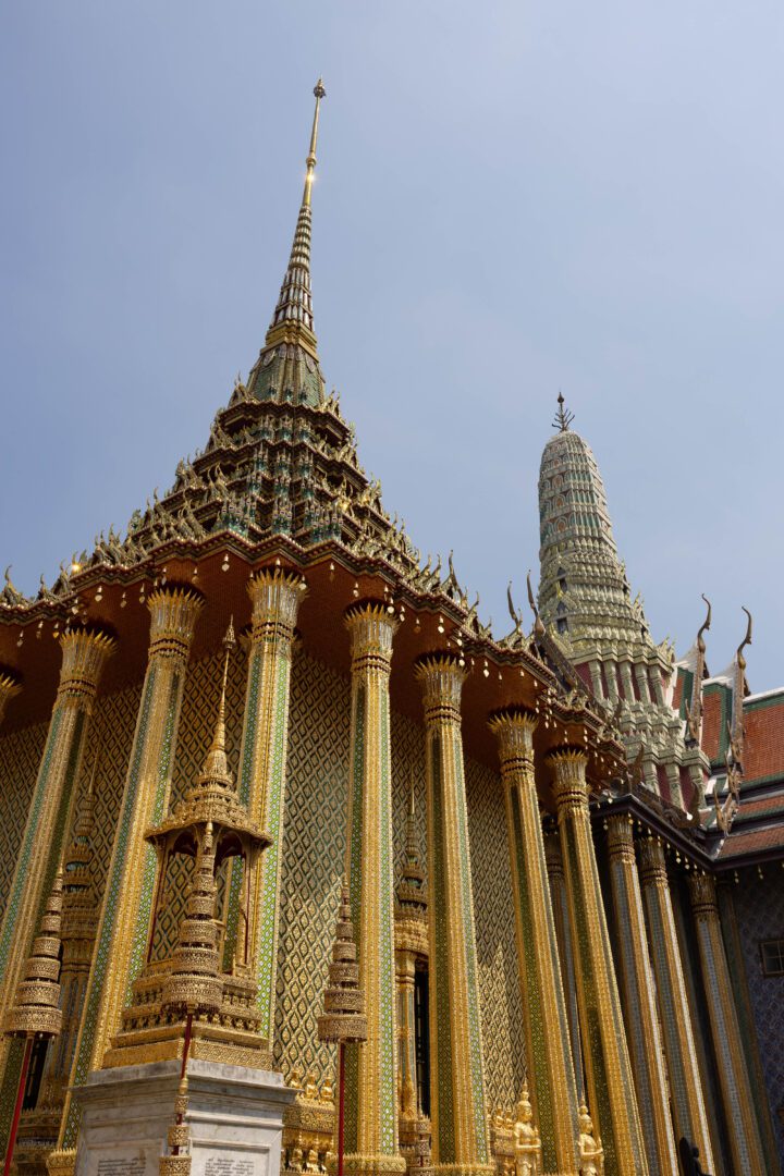 Golden spires and intricate mosaics adorn the exterior of a temple building at the Grand Palace in Bangkok, set against a clear blue sky.