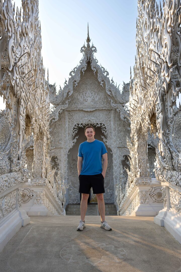 Man standing at the ornate entrance of Wat Rong Khun, the iconic White Temple in Chiang Rai, surrounded by detailed white sculptures and morning light.