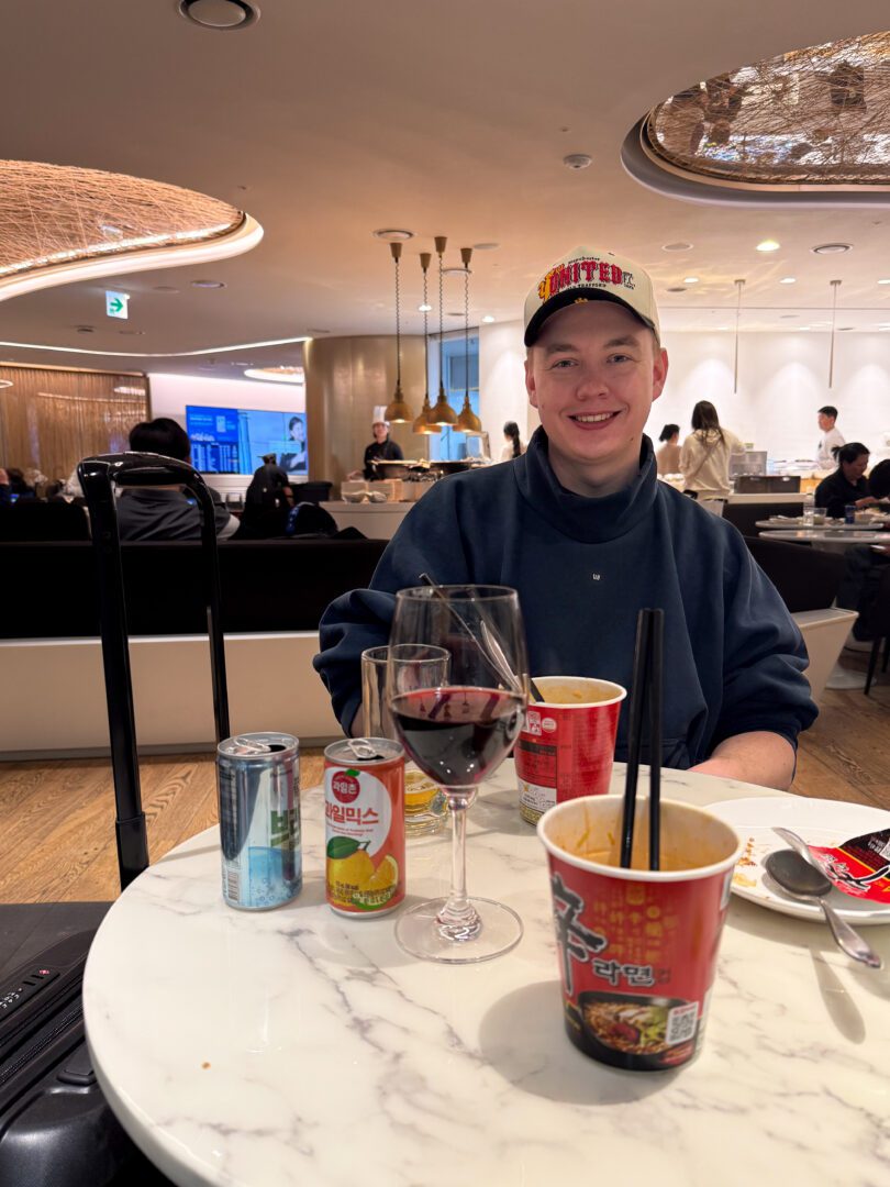 Man enjoying cup noodles, wine, and juice at a modern Korean airport lounge with warm lighting and a relaxed atmosphere before a flight.