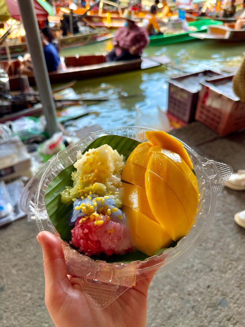 Colorful mango sticky rice served in a plastic container at Damnoen Saduak Floating Market, with boats and vendors in the background adding to the vibrant local atmosphere.