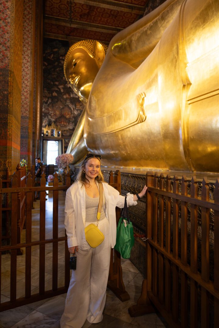 Woman posing beside the massive golden Reclining Buddha statue at Wat Pho in Bangkok, Thailand, surrounded by ornate temple decor.