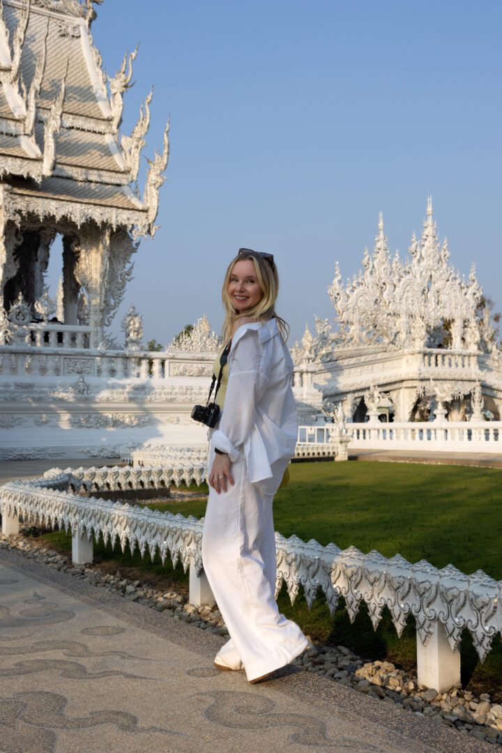 Woman in white outfit smiling in front of the intricate white carvings of Wat Rong Khun in Chiang Rai, Thailand, during golden hour.