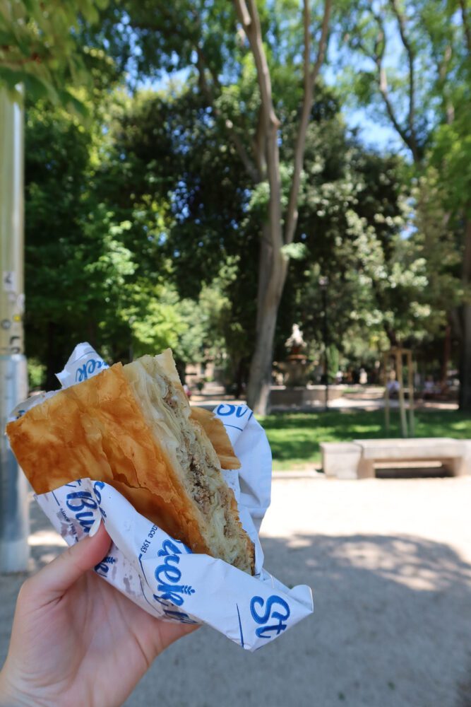Hand holding a savory meat burek from St Burek in Split, Croatia, wrapped in branded paper with a leafy park in the background—capturing a classic Balkan street food moment on a sunny day in Dalmatia.