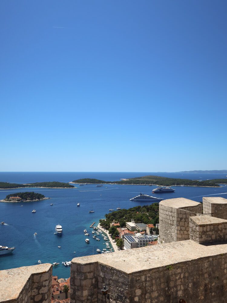 Panoramic view from the Fortica Fortress overlooking Hvar Town, Croatia, with yachts and sailboats scattered across the Adriatic Sea and the Pakleni Islands visible in the distance under a clear blue sky.