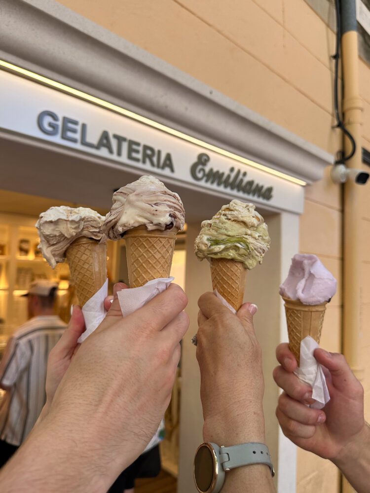 Four hands holding cones of artisan gelato in front of Gelateria Emiliana in Split, Croatia—showcasing creamy scoops in flavors like pistachio, stracciatella, and berry, perfect for a summer treat in the Old Town.