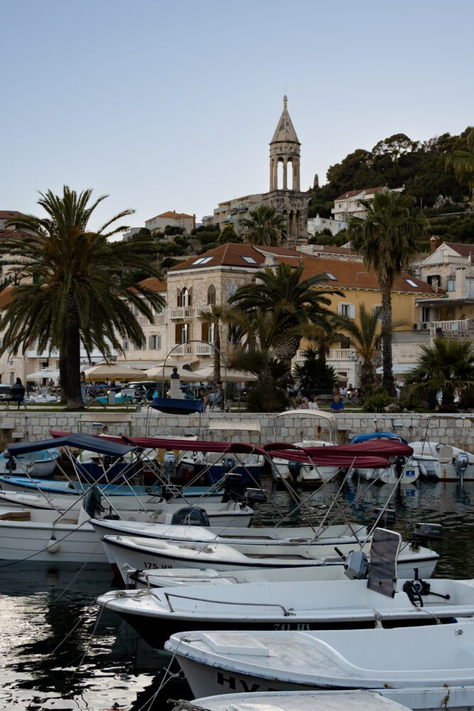 Boats docked along the Hvar harbor in Croatia, with palm trees, historic stone buildings, and the bell tower of the Franciscan Monastery rising in the background—capturing the relaxed charm of this Dalmatian island town at dusk.