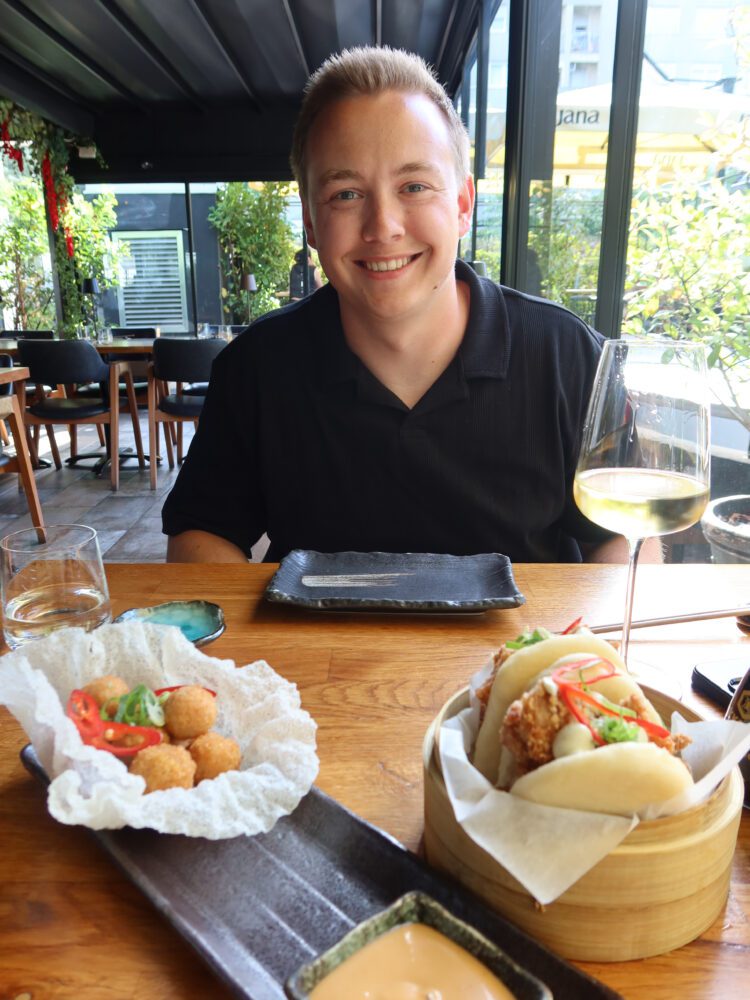 Man smiling at a modern Asian fusion restaurant in Zagreb with bao buns, croquettes, and white wine on the table