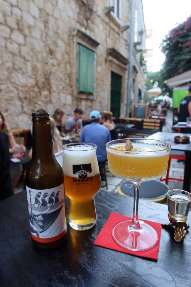 Craft beer and a citrus cocktail served at Lola Bar in Hvar, Croatia, with a Brewville Neretva Valley beer glass and a flower-garnished drink set on a black table along a charming stone alleyway filled with diners.