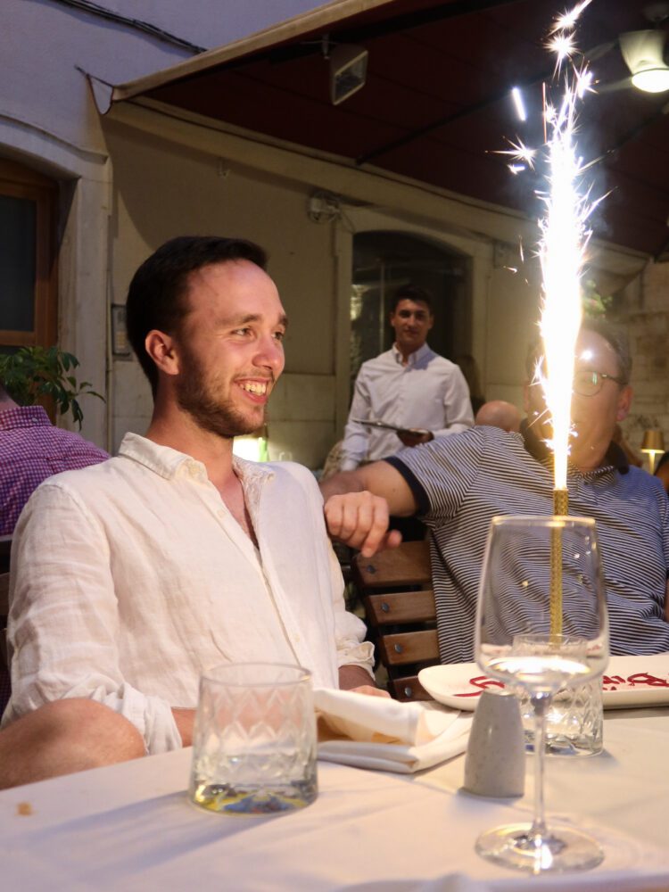 Luke smiling as a sparkling birthday candle lights up his dessert plate at a cozy outdoor restaurant in Split, Croatia—capturing a joyful moment during a summer evening celebration with friends.