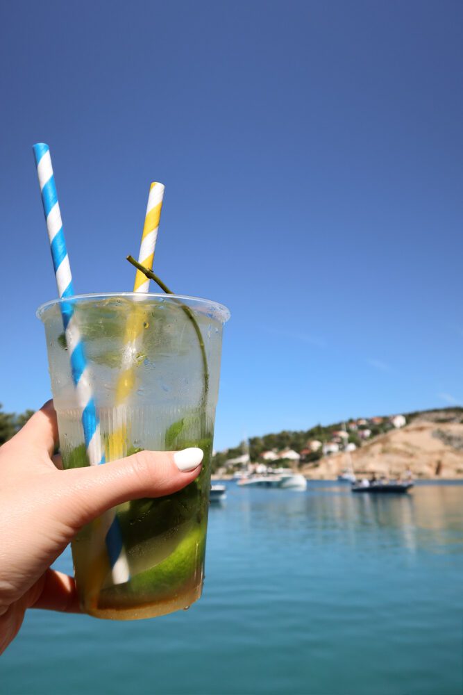 Close-up of a hand holding a fresh mojito in a plastic cup with blue and yellow striped straws, set against the turquoise waters and anchored boats of the Blue Lagoon near Split, Croatia, on a sunny summer day.