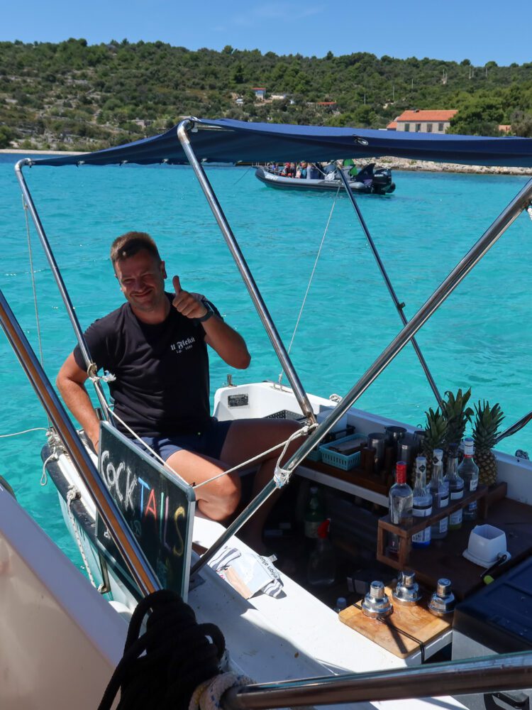 Smiling man giving a thumbs up from a floating cocktail boat at the Blue Lagoon near Split, Croatia, with bottles of liquor and pineapples arranged on deck—offering fresh mojitos on the turquoise Adriatic Sea.