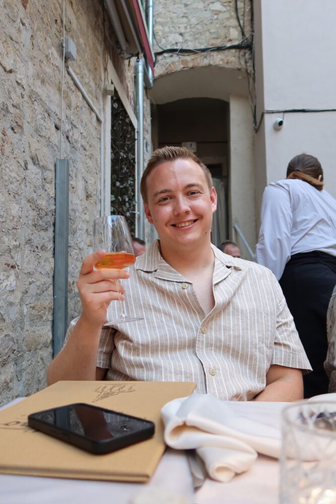 Ben smiling with a glass of orange wine at an outdoor table at Portofino restaurant in Split, Croatia, surrounded by stone walls and cozy alleyway ambiance—capturing the charm of a summer evening dining experience in the Old Town.