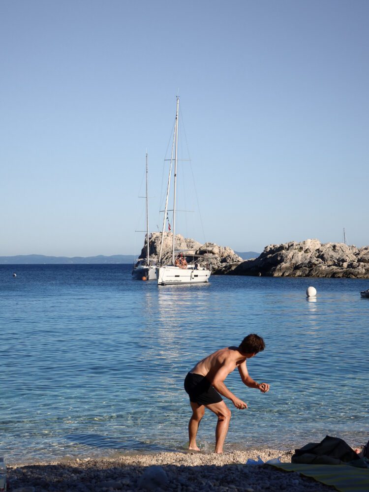 Man skipping rocks into the clear blue water at Zaraće Beach on Hvar Island, Croatia, with a sailboat anchored near rocky outcrops in the background—capturing a peaceful summer moment along the Adriatic coast.