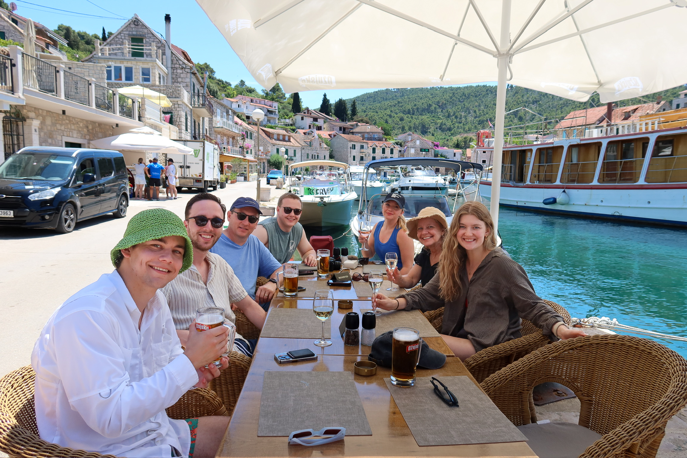 Group of friends and family enjoying drinks and lunch at a waterfront restaurant on Šolta Island, Croatia—seated under a large umbrella with boats docked behind them and colorful stone houses lining the harbor on a sunny summer day.