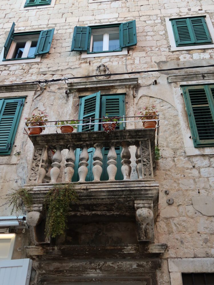 Charming stone building in Split, Croatia, featuring a weathered balcony with potted plants and green wooden shutters on all windows.