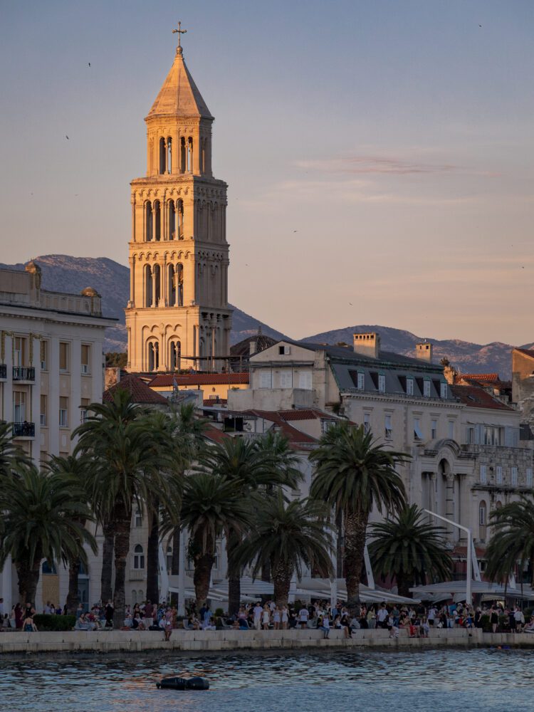 Saint Domnius bell tower glowing in the warm golden hour light above the Riva promenade in Split, Croatia, with palm trees, historic buildings, and crowds lining the waterfront as the sun sets over the Adriatic.