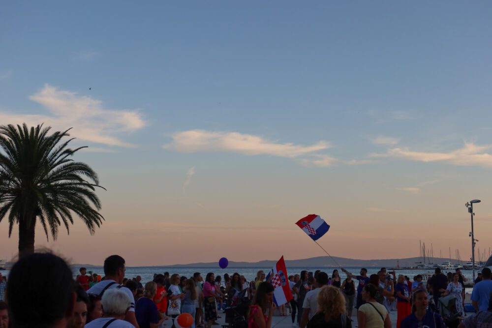 Crowd gathered along the Split, Croatia waterfront at sunset, with Croatian flags waving and sailboats docked in the background under a colorful evening sky.