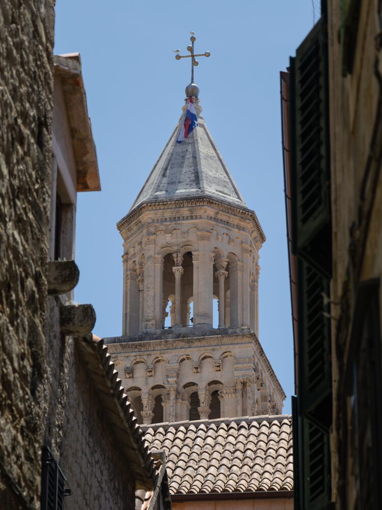 Close-up view of the bell tower of Saint Domnius Cathedral in Split, Croatia, framed between narrow stone alleyways and traditional terracotta rooftops—highlighting the Romanesque architecture and historic charm of Diocletian’s Palace.
