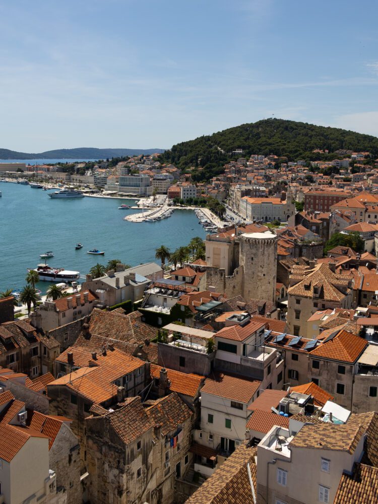 Panoramic view of Split, Croatia, featuring the terracotta rooftops of the Old Town, the Adriatic Sea lined with yachts and palm trees, and Marjan Hill rising in the background—captured from the top of Saint Domnius bell tower.