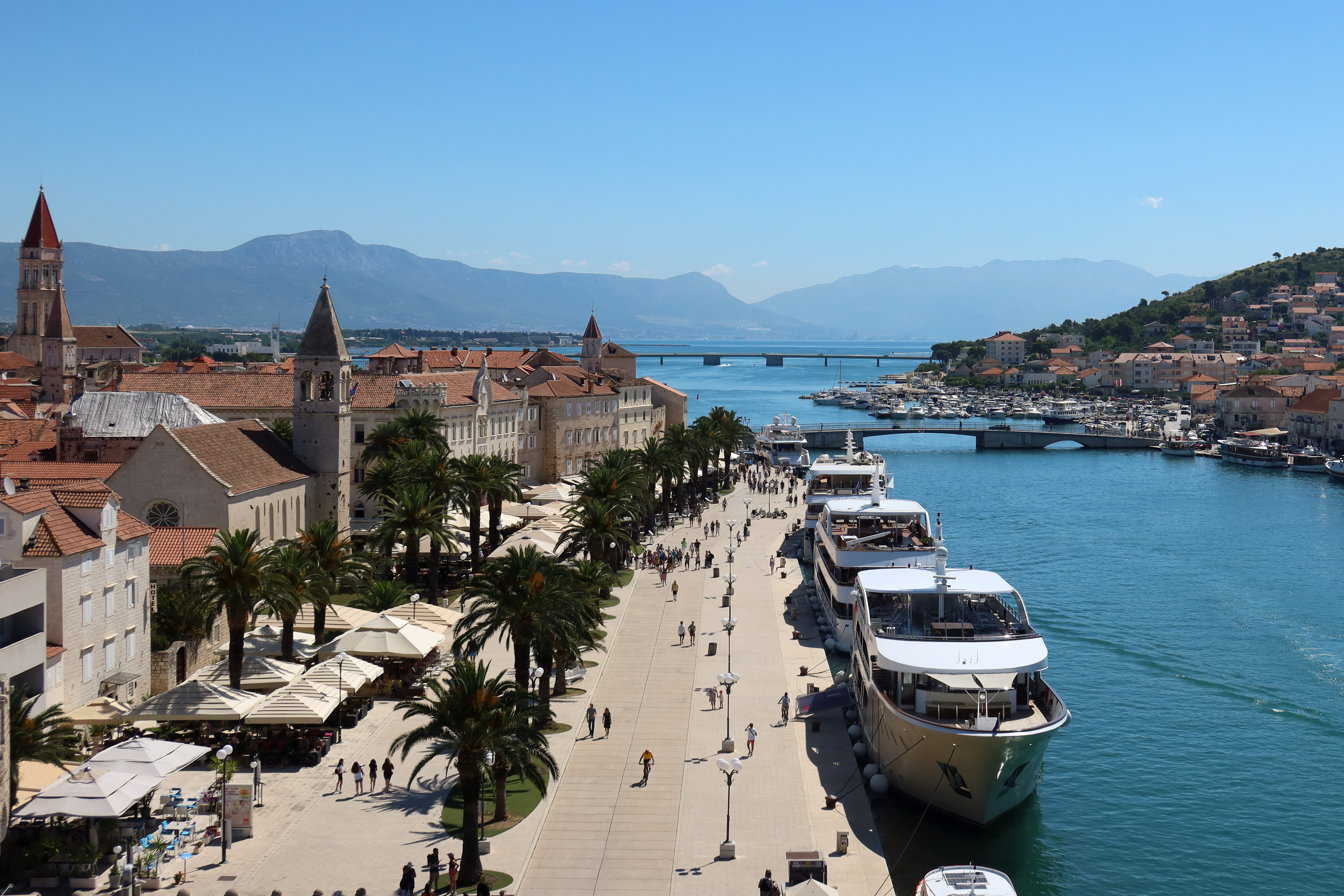 Aerial view of the Trogir waterfront promenade in Croatia, lined with palm trees, yachts, and medieval stone buildings with red-tiled roofs—set against a backdrop of mountains and the Adriatic Sea on a clear summer day.