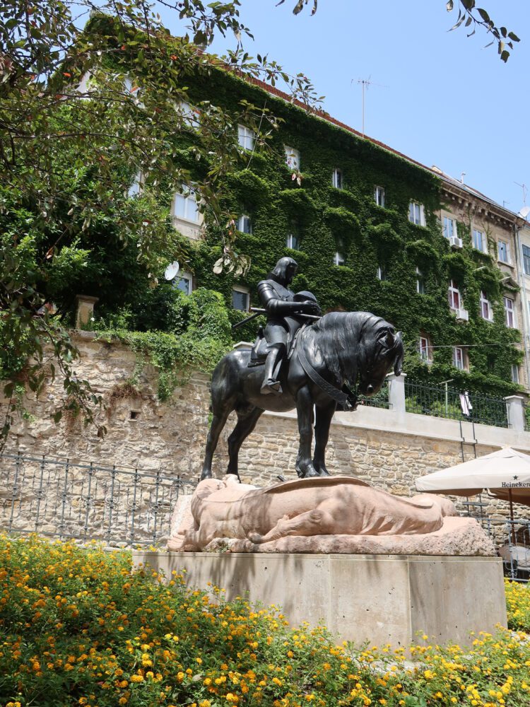 Equestrian statue of St. George slaying the dragon in Zagreb, Croatia, set against a lush ivy-covered building and vibrant yellow flowers on a sunny day