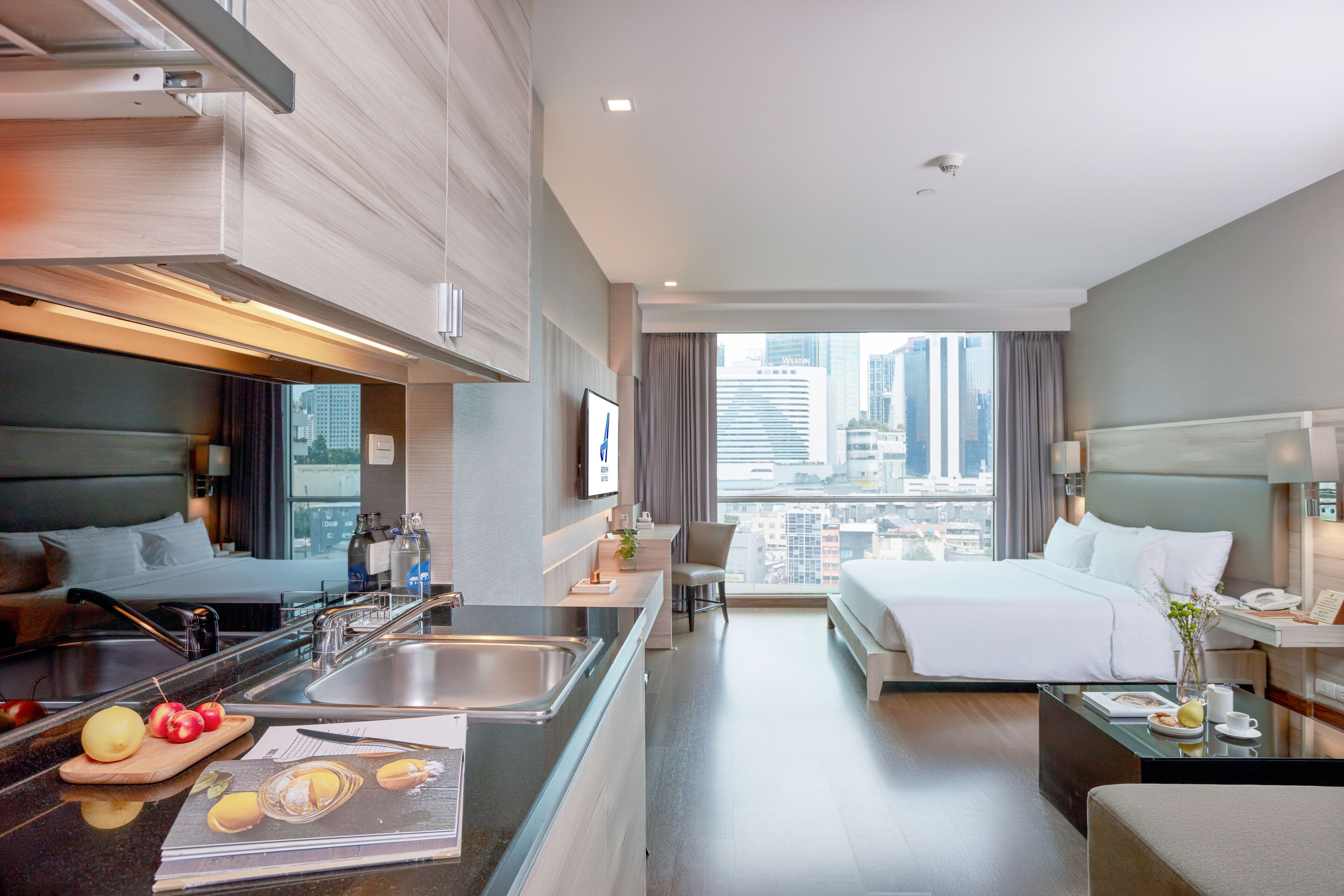 Modern studio at Adelphi Suites Bangkok featuring a compact kitchenette, cozy seating area, and floor-to-ceiling windows with sweeping views of the city skyline.