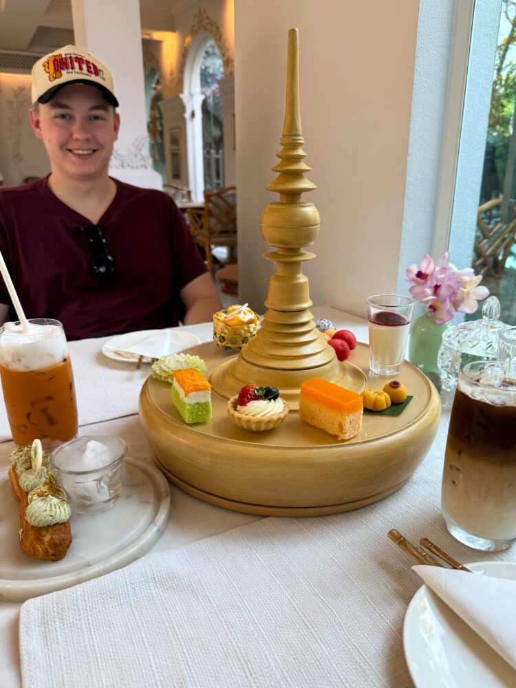 Elegant Thai afternoon tea set at The Inside House in Chiang Mai, Thailand, featuring traditional and modern desserts served on a golden tiered platter, with iced coffee and Thai tea in a luxurious colonial-style setting.
