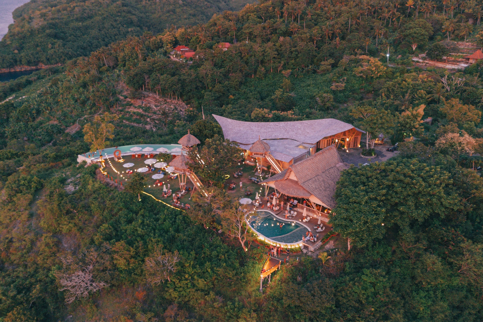 Aerial view of Amok Sunset in Nusa Penida, a clifftop bamboo restaurant and bar with glowing lights, sunset views, and a jungle backdrop overlooking the sea.