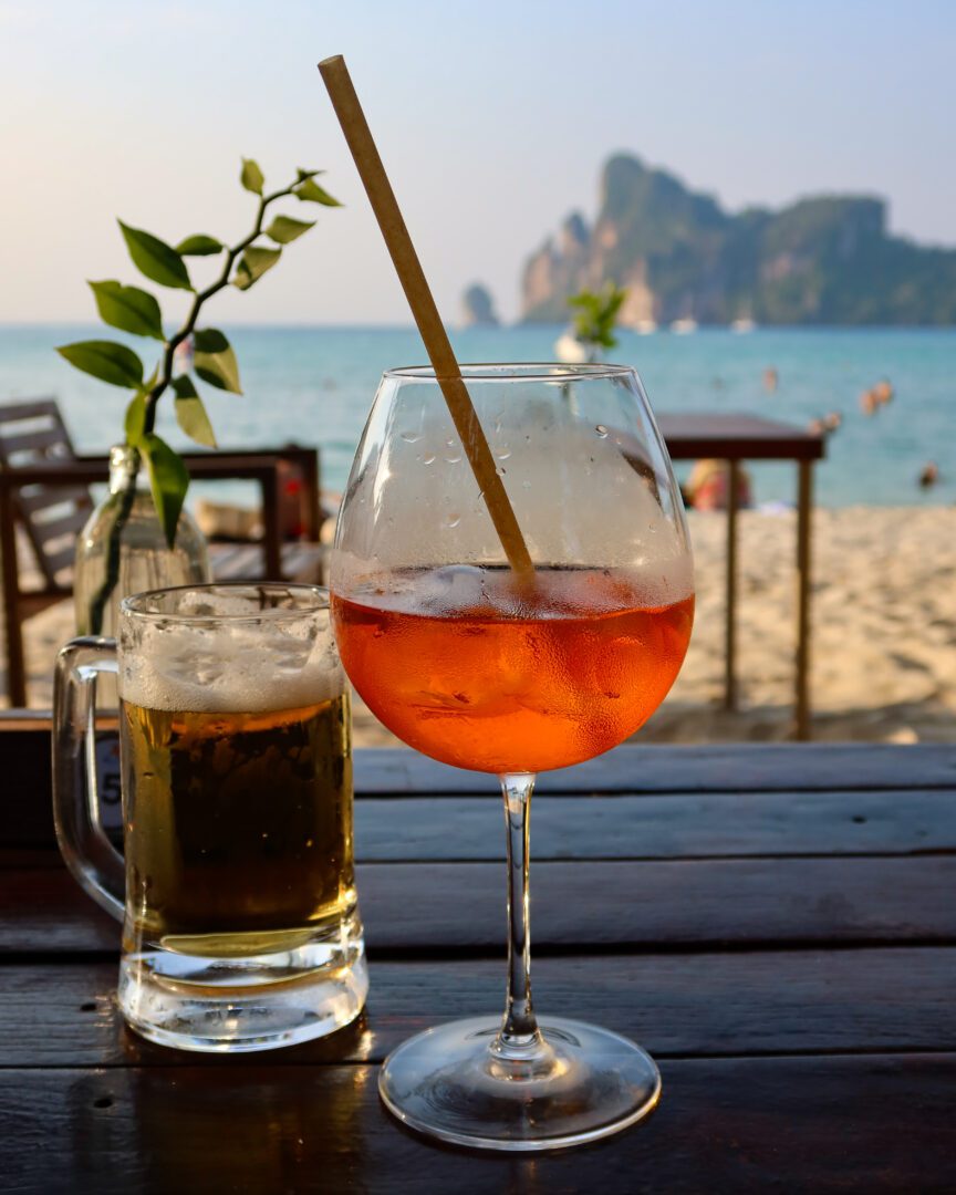 Refreshing Aperol Spritz and cold beer on a wooden table with a beachfront view of Koh Phi Phi’s limestone cliffs and turquoise water in the background.