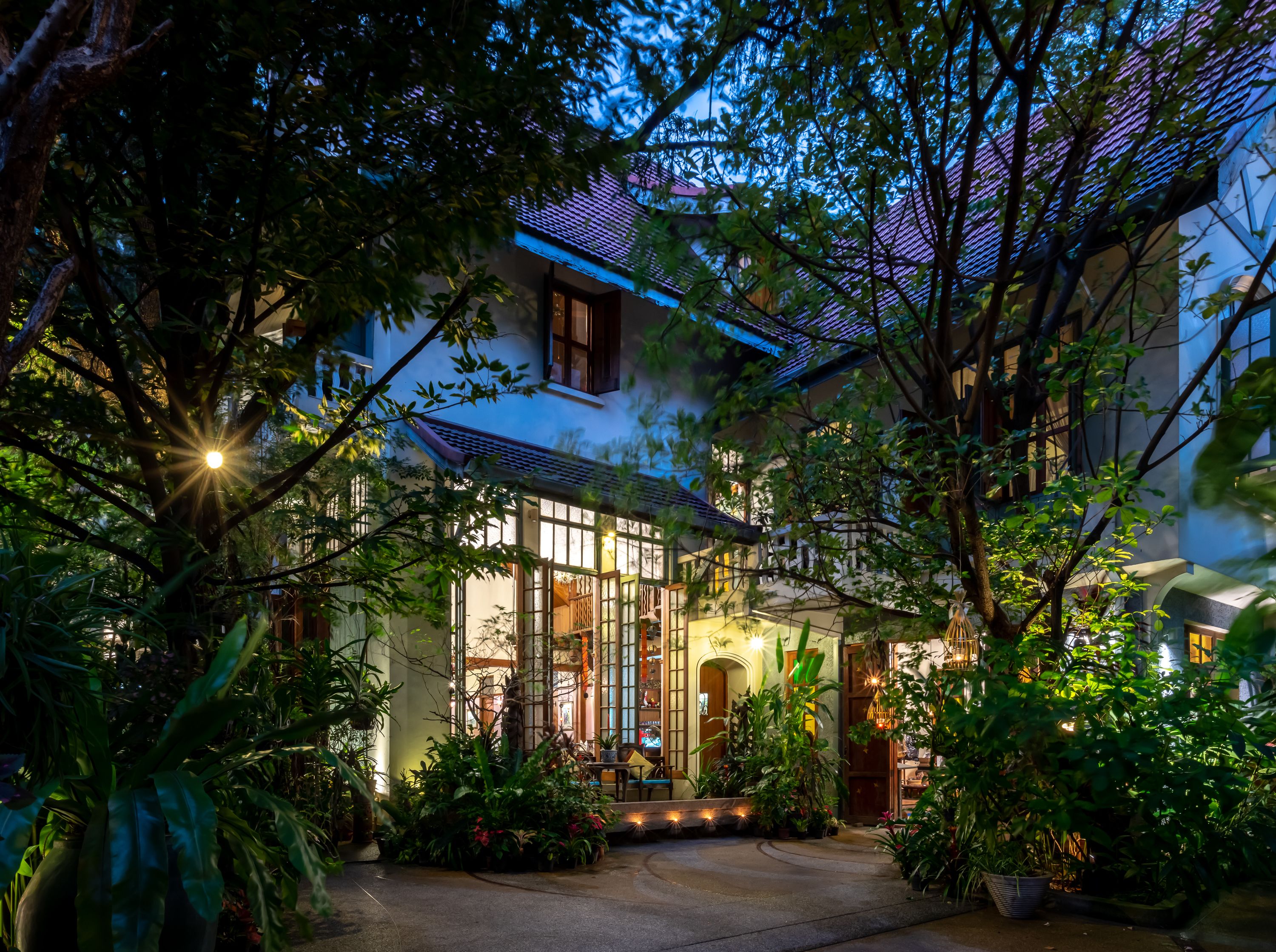 Charming evening view of Ariyasomvilla’s leafy entrance, where warm lights and lush tropical greenery frame the elegant, colonial-style boutique hotel in central Bangkok.
