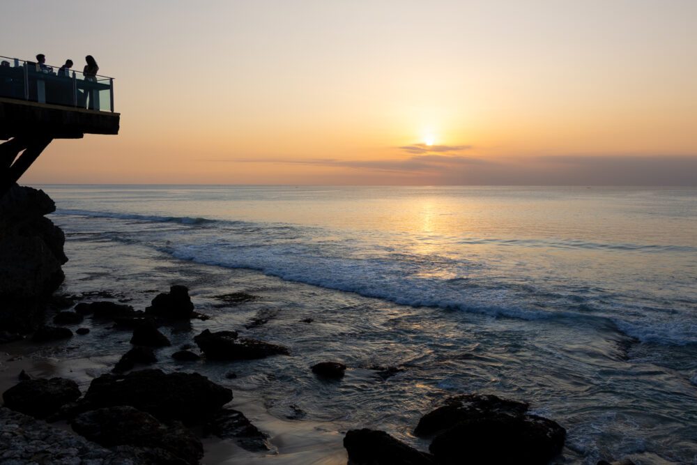 Golden sunset view from Ayana’s Rock Bar in Bali, with silhouettes of guests enjoying cocktails perched above the crashing waves of Jimbaran Bay.