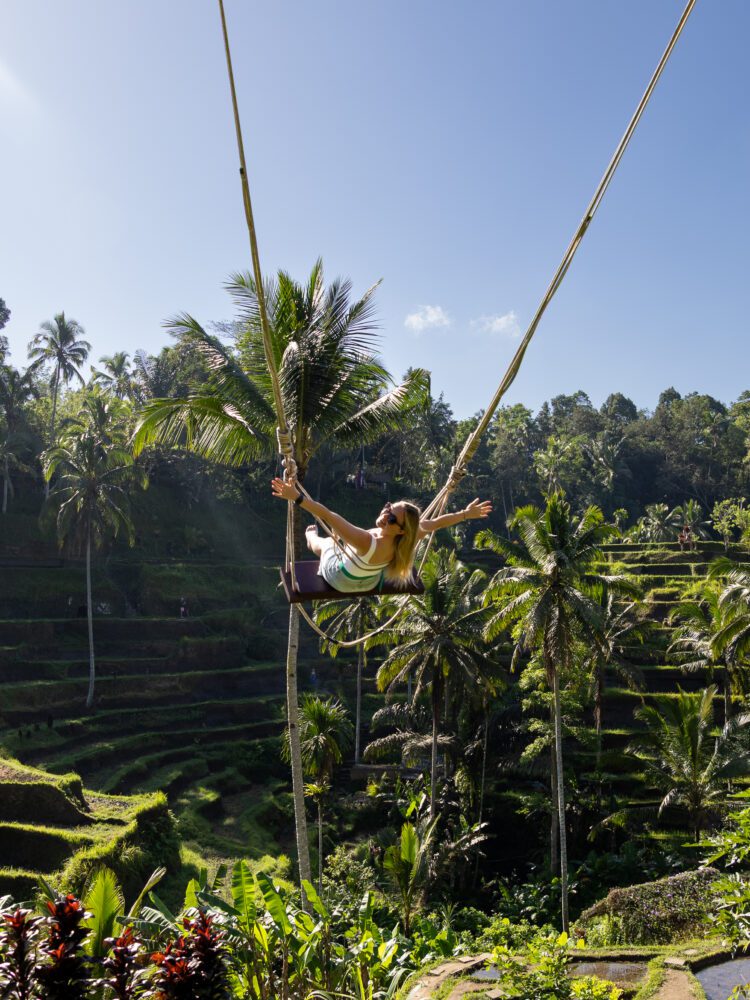 Woman soaring on the iconic Bali Swing above the lush Tegalalang Rice Terraces, surrounded by tropical palm trees and dramatic green landscapes under a clear blue sky.
