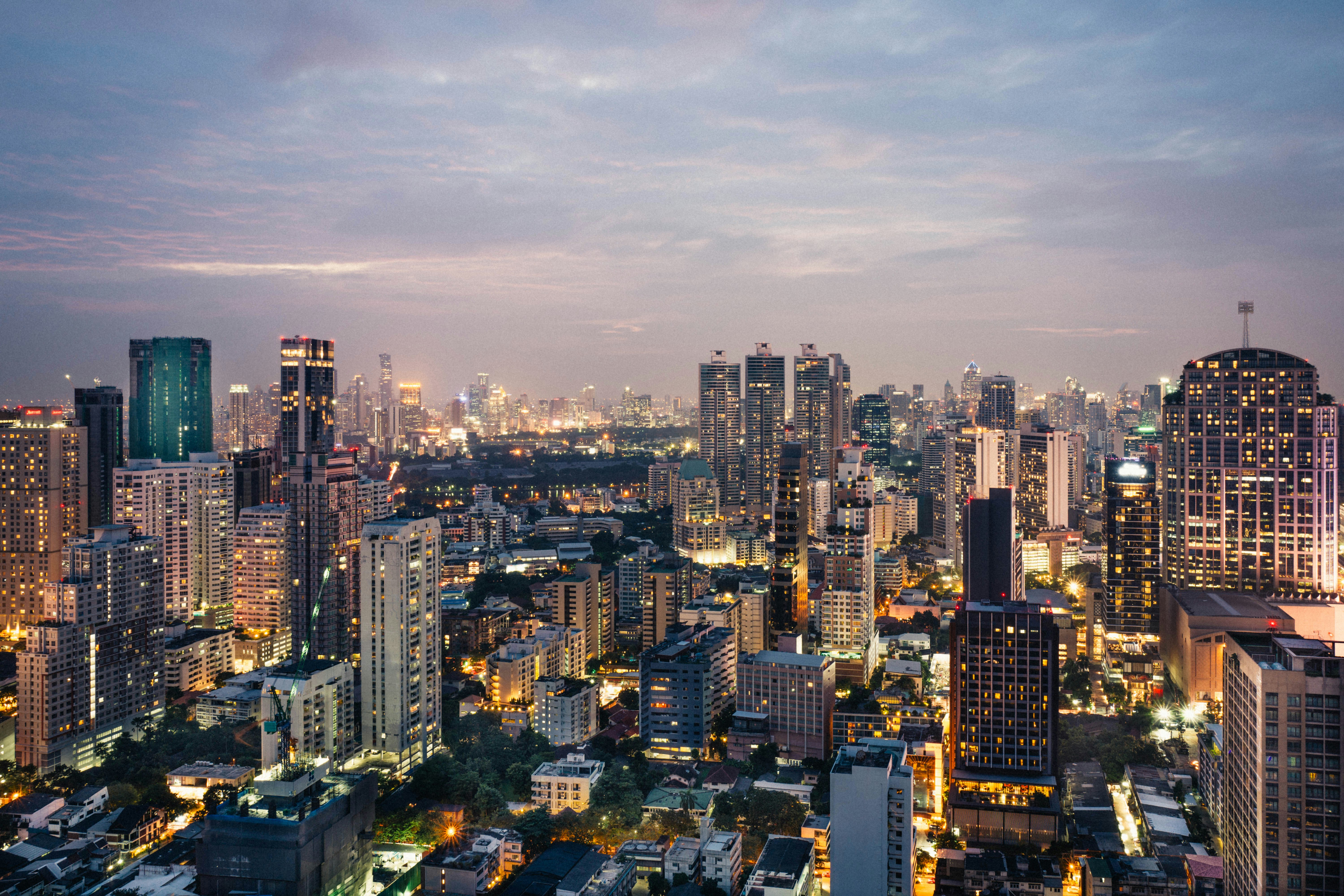 Bangkok’s downtown skyline glows at dusk, with high-rise buildings and city lights stretching toward the horizon under a soft, cloud-streaked sky.