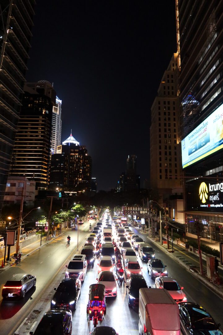 Streams of headlights and taillights fill a major road in Bangkok, capturing the city's infamous nighttime traffic against a backdrop of illuminated skyscrapers.
