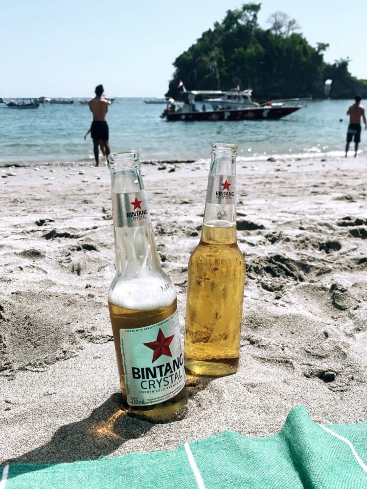 Chilled Bintang Crystal beers resting in the sand at Crystal Bay Beach, Nusa Penida, with boats bobbing in the turquoise water and a lush island backdrop.