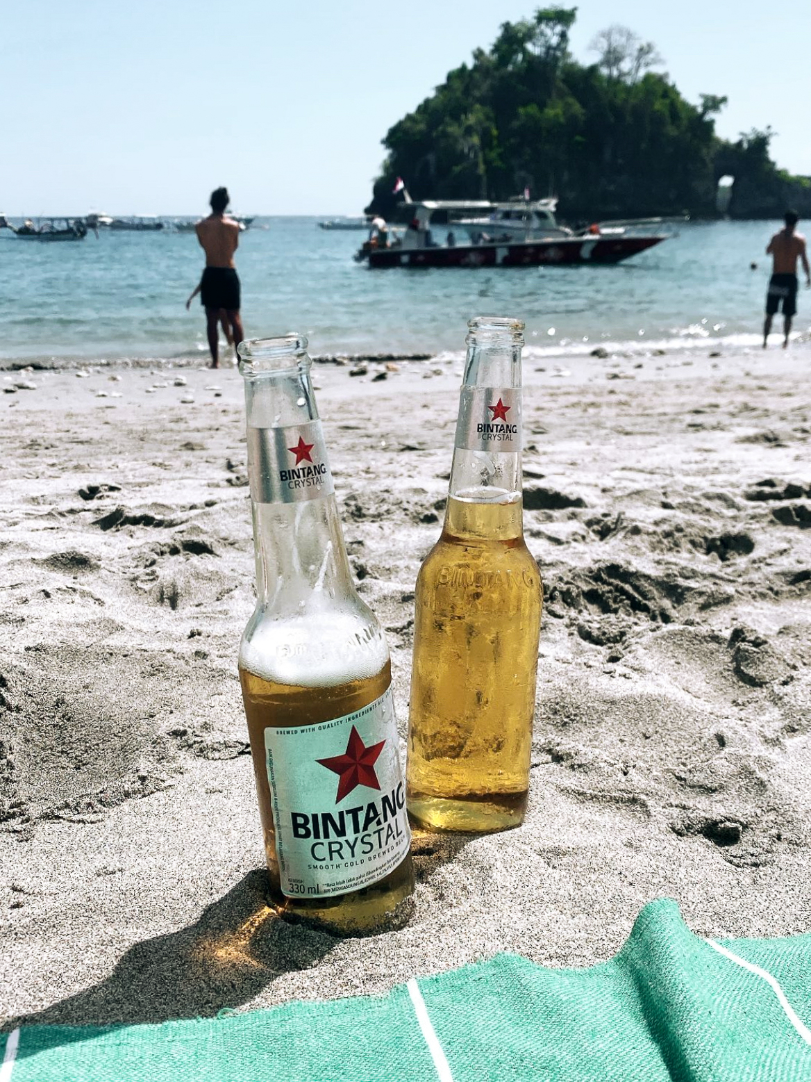 Chilled Bintang Crystal beers resting in the sand at Crystal Bay Beach, Nusa Penida, with boats bobbing in the turquoise water and a lush island backdrop.