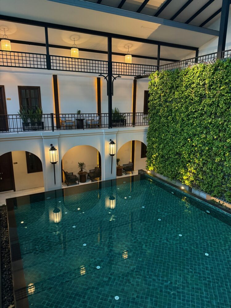 Serene indoor pool at Makkha Spa in Chiang Mai, Thailand, featuring emerald-green tiles, a lush vertical garden wall, and elegant colonial-style architecture with ambient lighting and private seating areas.
