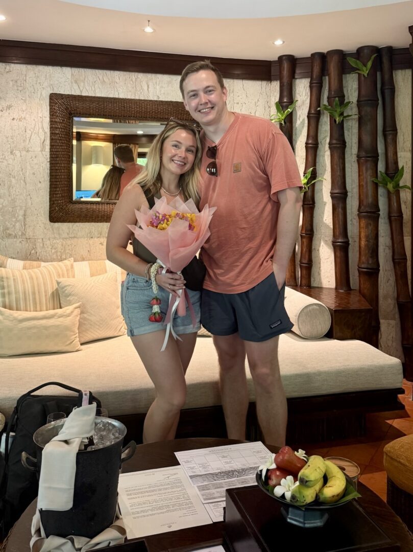 Couple smiles during check-in at Rayavadee Resort, holding a welcome bouquet of tropical flowers with fresh fruit and champagne awaiting in their luxurious villa.