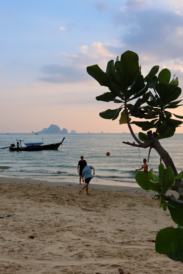Group of boys playing soccer on Ao Nang Beach in Krabi, Thailand at sunset, with a traditional longtail boat and distant islands in the background.