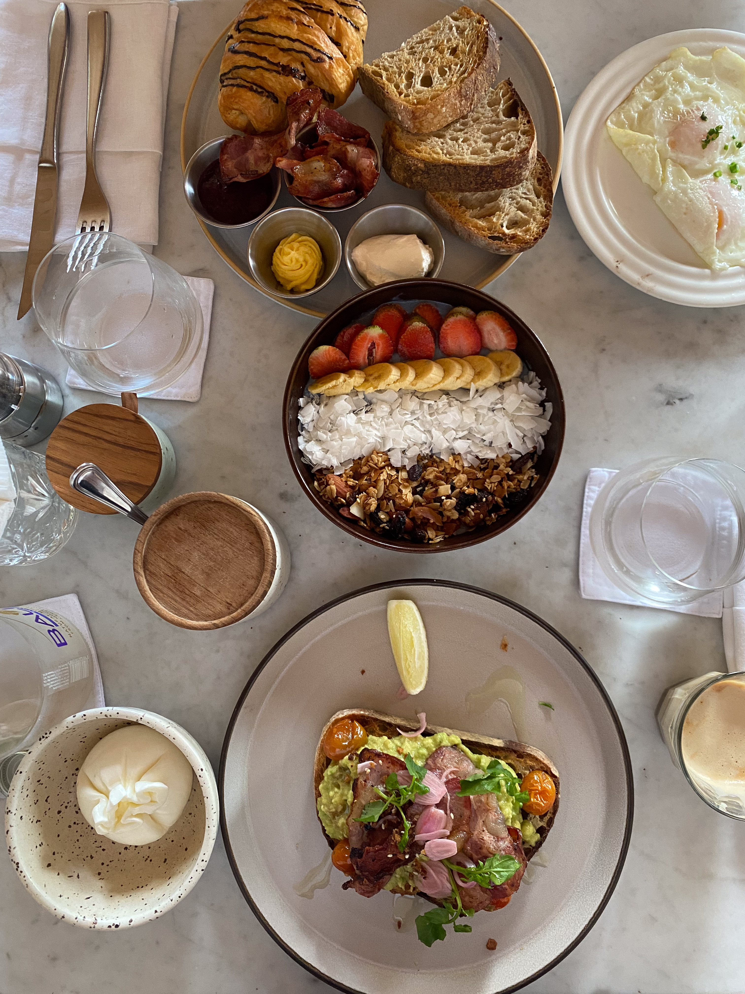 Delicious brunch spread at Gooseberry Restaurant in Uluwatu, featuring avocado toast with bacon, smoothie bowl topped with fresh fruit and granola, pastries, and eggs, all beautifully arranged on a marble table.