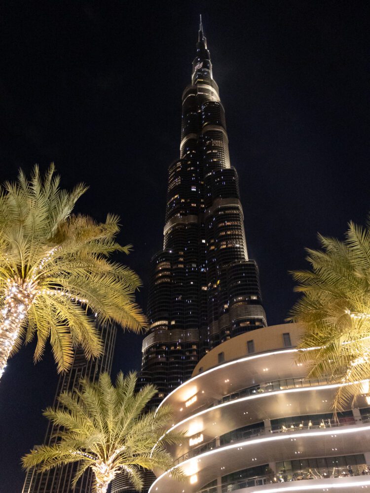 The Burj Khalifa illuminated at night during a light show, rising above palm trees and the Dubai Mall’s curved architecture in a dazzling display of modern design and luxury.