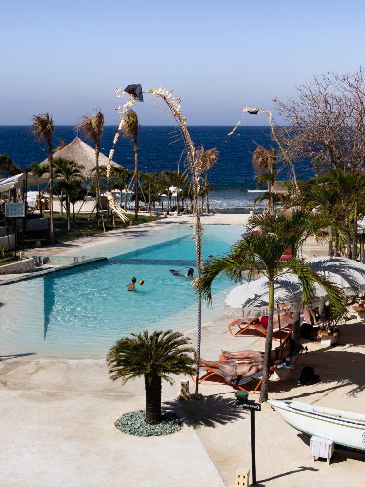 Beachside pool at Cactus Day Club in Nusa Penida, with ocean views, palm trees, festive penjor decorations, and guests lounging under umbrellas on bright orange sunbeds.