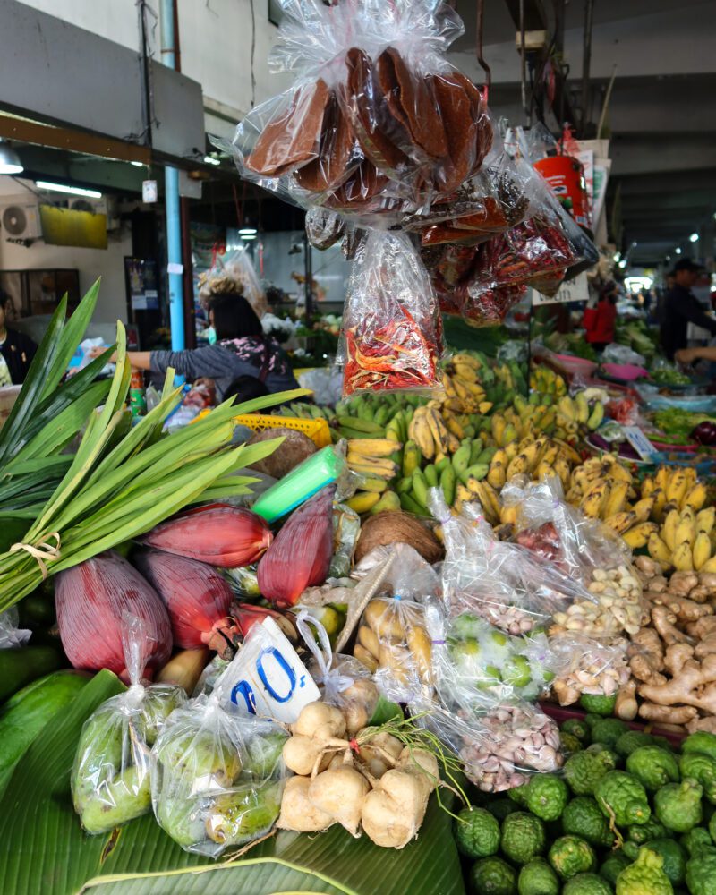 Colorful fresh produce and spices on display at a bustling local market in Chiang Mai, Thailand, featuring tropical fruits, herbs, and vegetables like banana flowers, kaffir limes, ginger, and chili peppers.