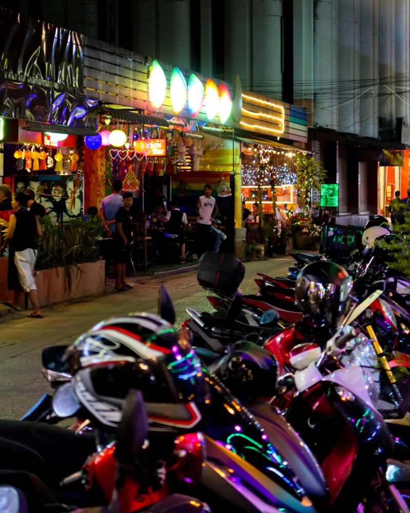 Colorful motorbikes line a lively street outside a vibrant bar near Chiang Mai Night Market, Thailand, glowing with neon lights and rainbow décor as locals and tourists gather to enjoy the nightlife.