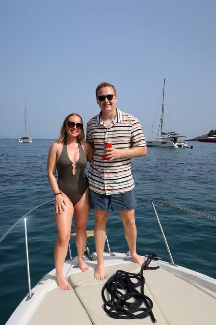Couple standing barefoot on a boat deck, smiling with drinks in hand, surrounded by anchored yachts near the Elaphiti Islands.