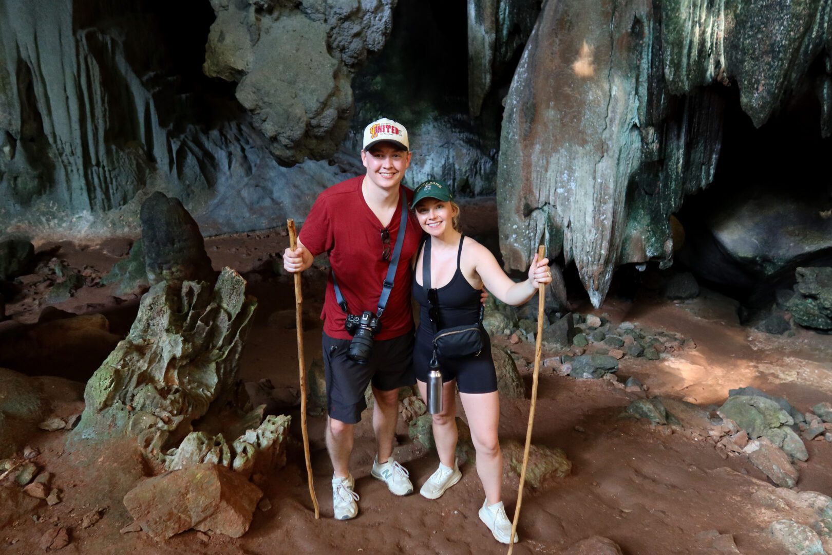 A couple poses with walking sticks inside a cave in Khao Sok National Park, surrounded by striking limestone formations and earthy red terrain.