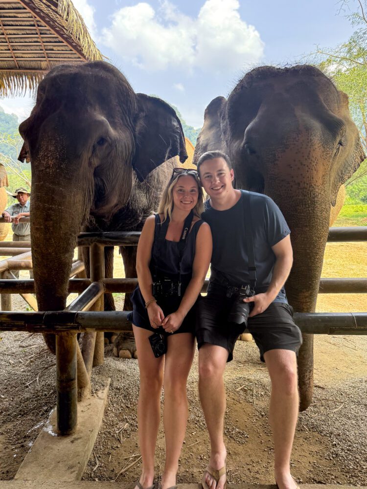 Smiling couple posing with two rescued elephants at an ethical elephant sanctuary in Thailand, highlighting responsible wildlife tourism and up-close animal encounters in a natural setting.
