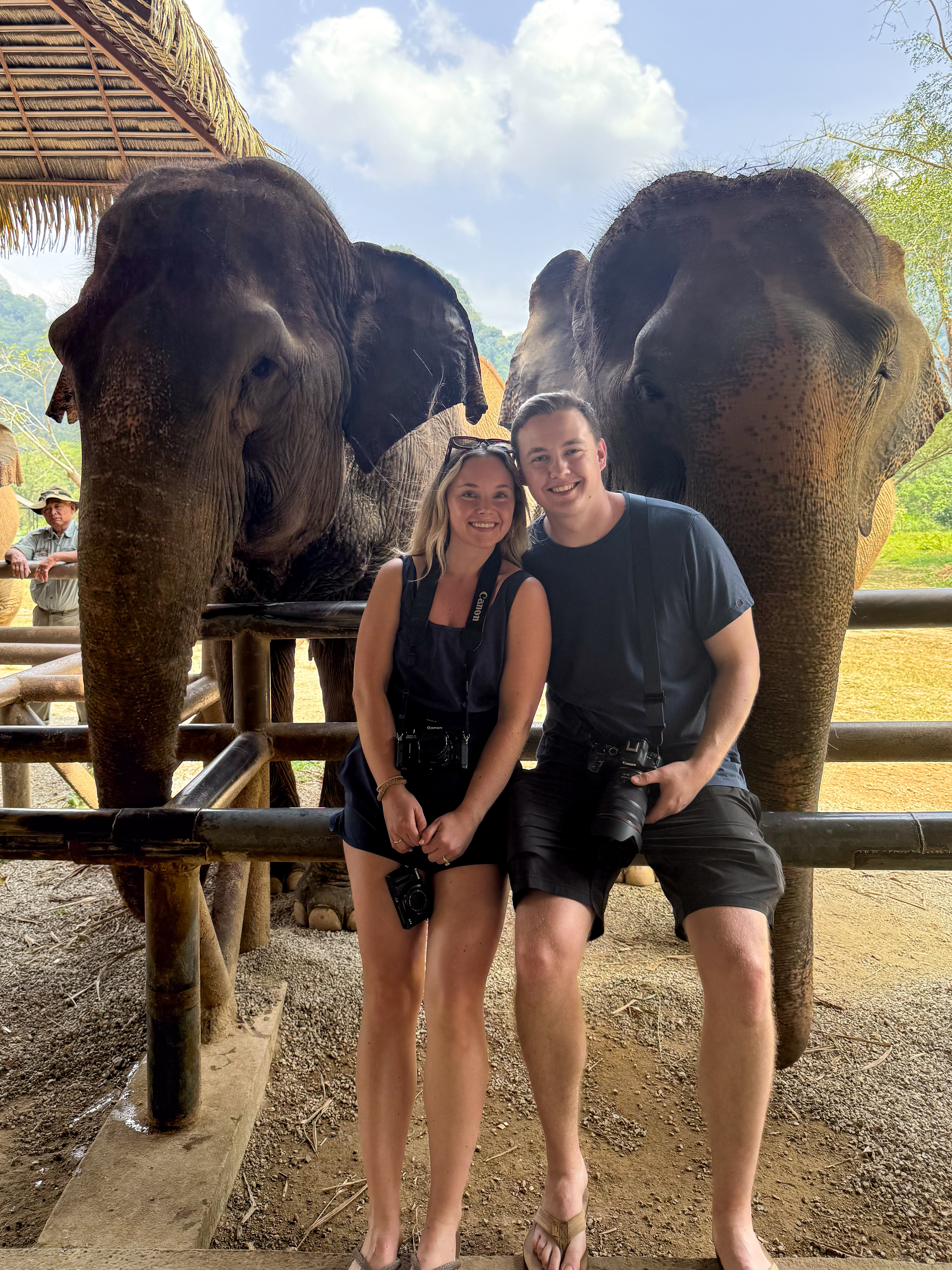 Smiling couple posing with two rescued elephants at an ethical elephant sanctuary in Thailand, highlighting responsible wildlife tourism and up-close animal encounters in a natural setting.