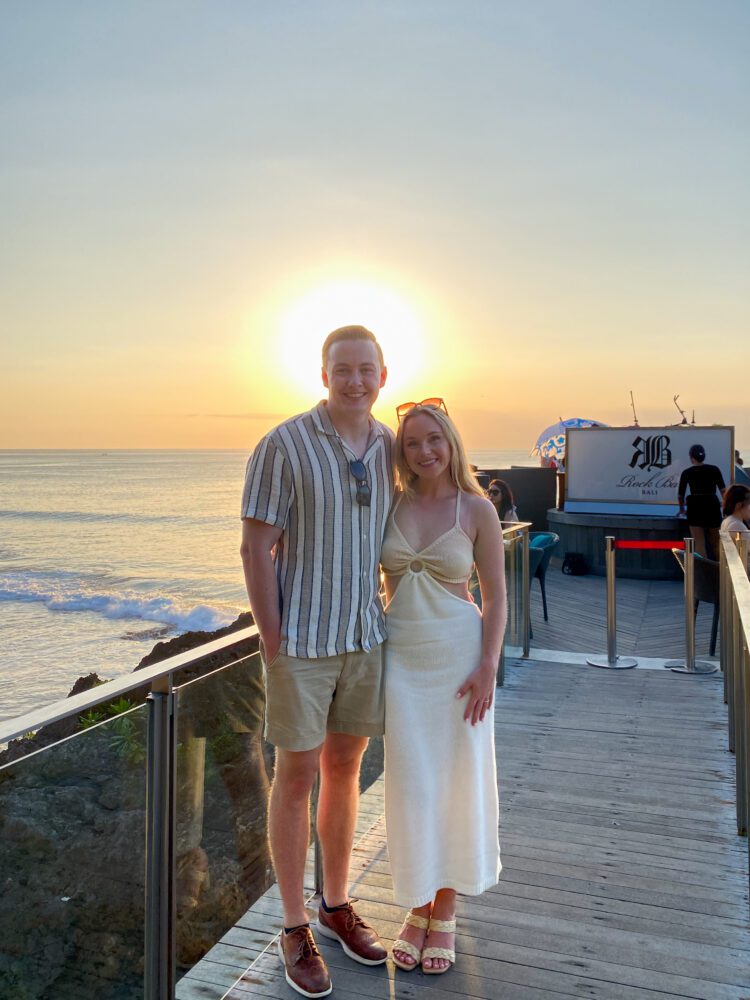 Couple standing on a boardwalk at Rock Bar Uluwatu with the sun setting directly behind them and the Indian Ocean glowing in golden light.